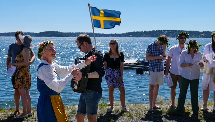 Allsång vid Lerviks brygga. En kvinna i svensk landskapsdräkt och blomkrans i håret leder allsång tillsammans med en man med dragspel. I vy mot fjärden i bakgrunden: sommarklädda ungdomar och en kvinna som håller upp en svensk flagga.